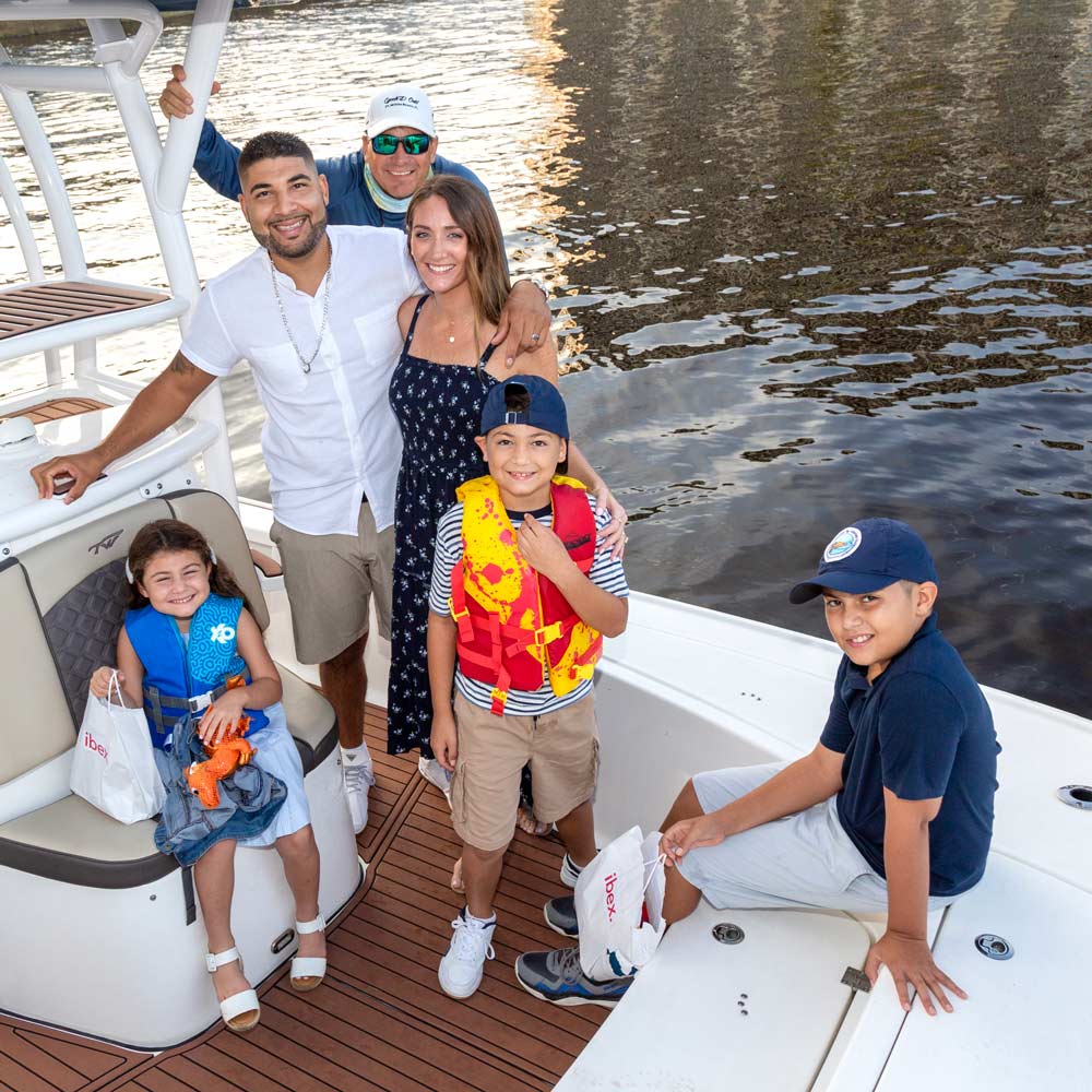 A little boy in a navy blue ball cap stands on a boat holding a fishing pole and smiling at the camera while a man in a white ball cap holds a small fish out to him.