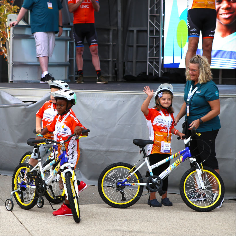 Three children with helmets and red, white, and orange UHCCF jerseys stand with their bicycles in front of a stage. One little girl stands next to a woman in a teal UHCCF polo shirt and waves to the crowd.