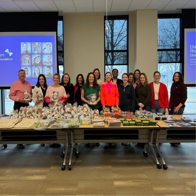 A group of people from the E&I People Team stands together behind a table full of Grins to Go activity bag items, including coloring books, pop-its, and stuffed animals.