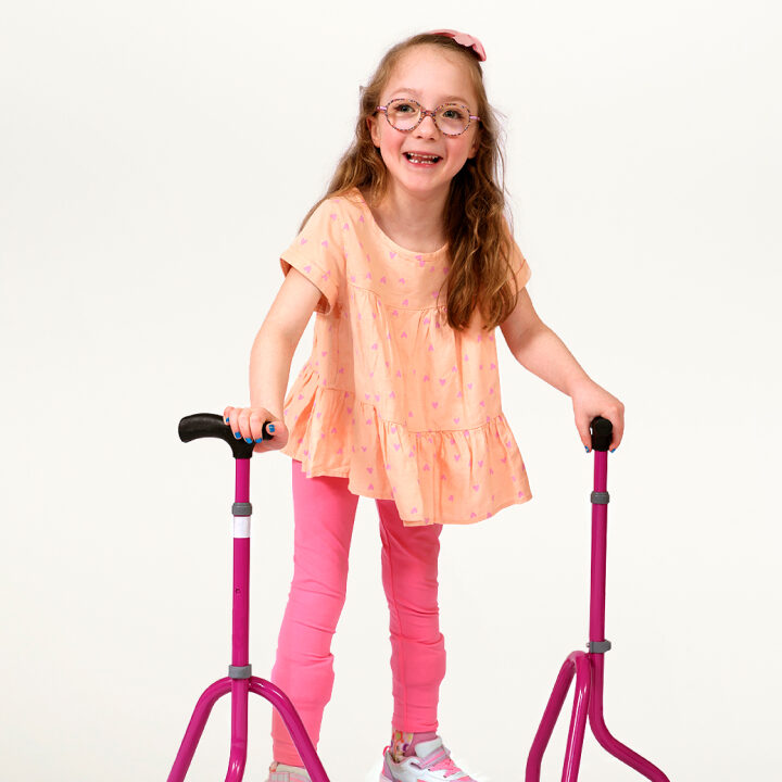 A little girl named Naomi stands with her pink walker and smiles at the camera.