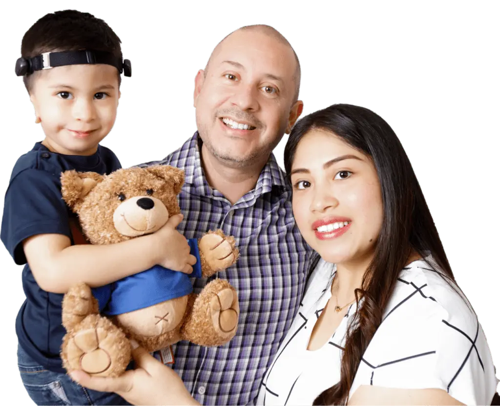A family smiles at the camera with their arms wrapped around each other. The dad holds the little boy, Adrian, from Houston, TX while he hugs an Oliver the Bear stuffed animal. The mom stands on the dad’s other side, holding Oliver’s foot.