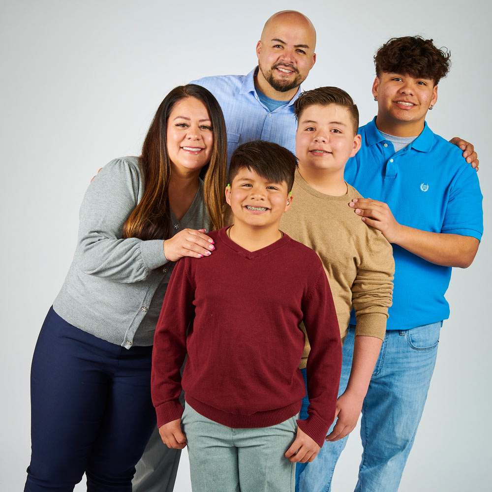 A little boy named Noah from Thornton, CO stands with his thumbs in his pockets. His family stands around him, smiling at the camera with their hands on each other’s shoulders.