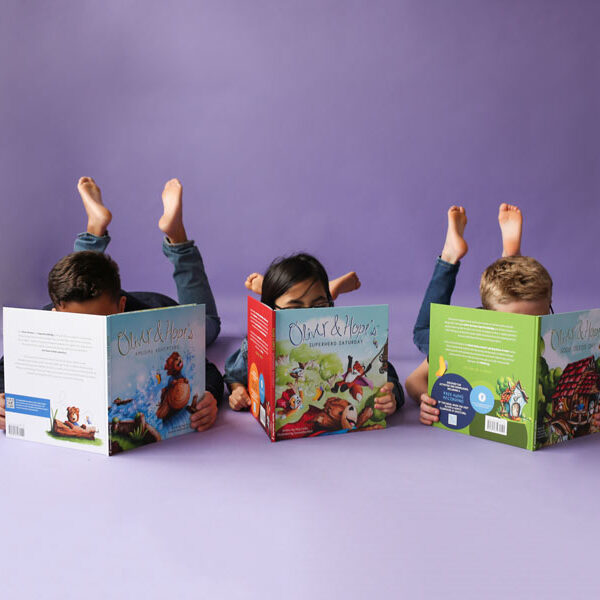 Three children lay on their tummies with their feet in the air, their faces hidden by open Oliver & Hope picture books.