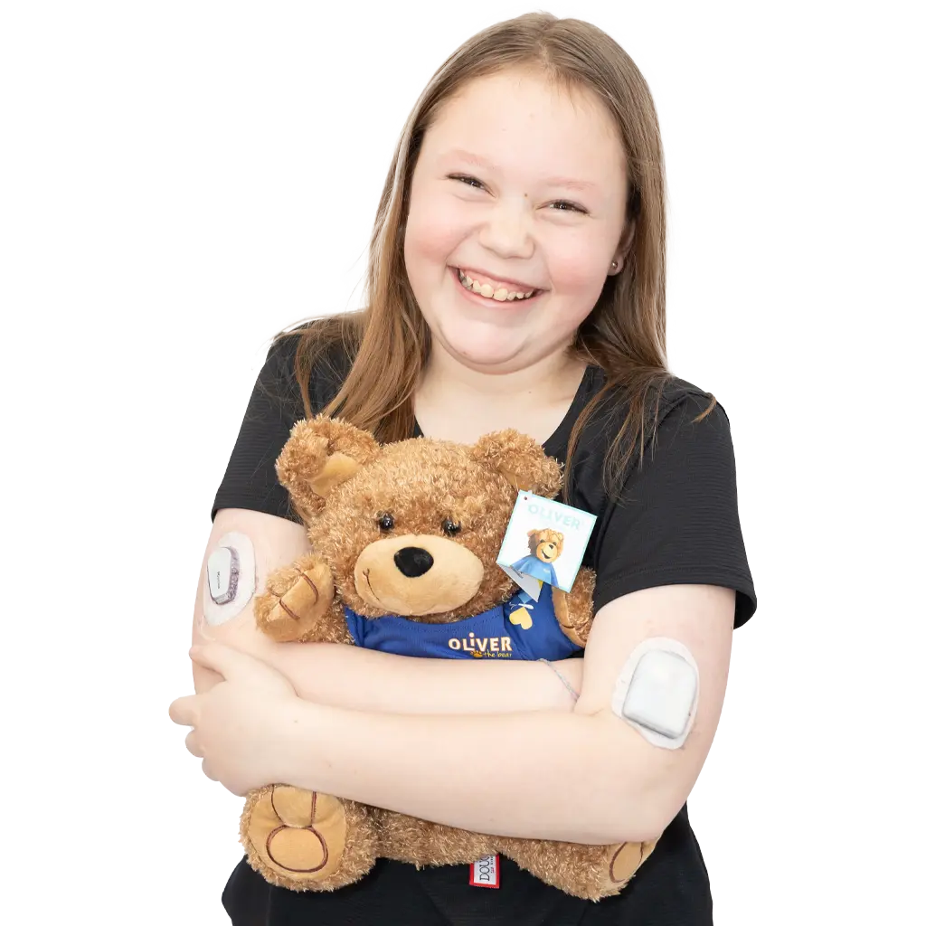 A little girl named Zayley smiles widely at the camera while hugging a stuffed Oliver the Bear.
