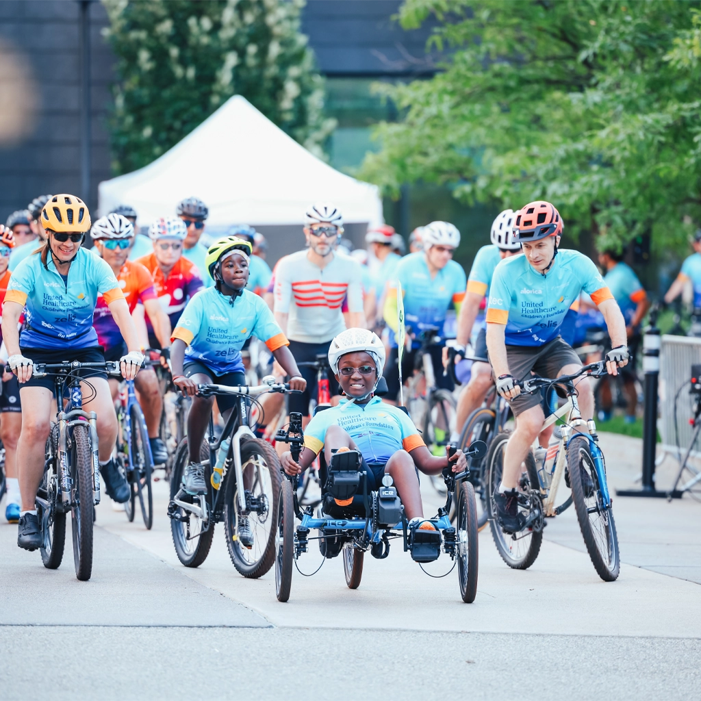 A group of people ride bicycles together at the Minnesota Century Ride event. The person at the front is riding a three-wheel wheelchair bicycle and smiling at the camera.