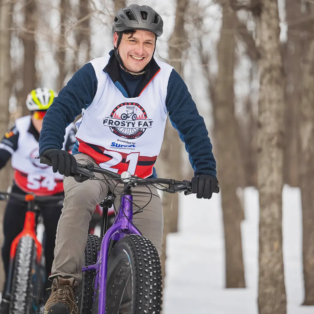 A man with a black helmet and an event shirt with the number 21 on it rides a purple bike through a forest at a Frosty Fat Tire event and smiles at the camera. There is snow on the ground. A person on a red bike with sunglasses, a yellow helmet, and an event shirt with the number 54 on it rides behind him.
