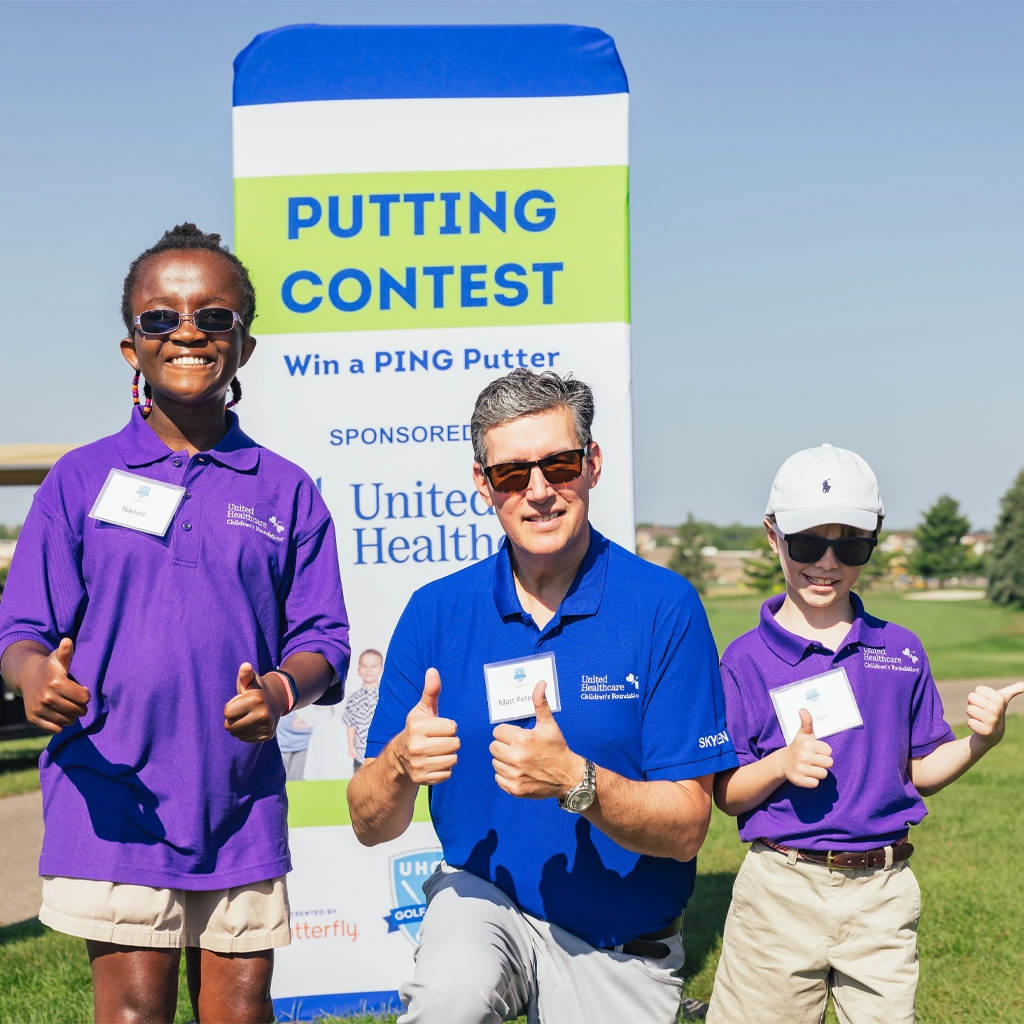 A little girl in sunglasses and a purple event shirt and a little boy also in a purple event shirt and sunglasses with a ball cap stand on either side of a man in a blue event shirt and sunglasses. The man is down on one knee and they are all smiling at the camera with both thumbs up.
