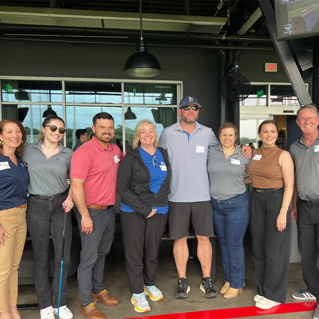 A group of eight men and women pose for a picture at a Louisiana Top Golf event. One woman is holding a blue golf club, and some have their arms around one another.