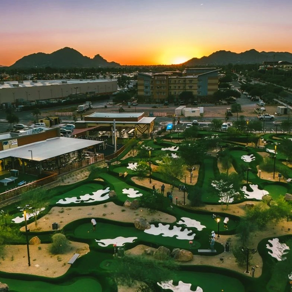 The sky glows purple, orange, and yellow as the sun sets behind the mountains at an Arizona Putt for Hope event. Several people are putting on the putt putt course under lights.