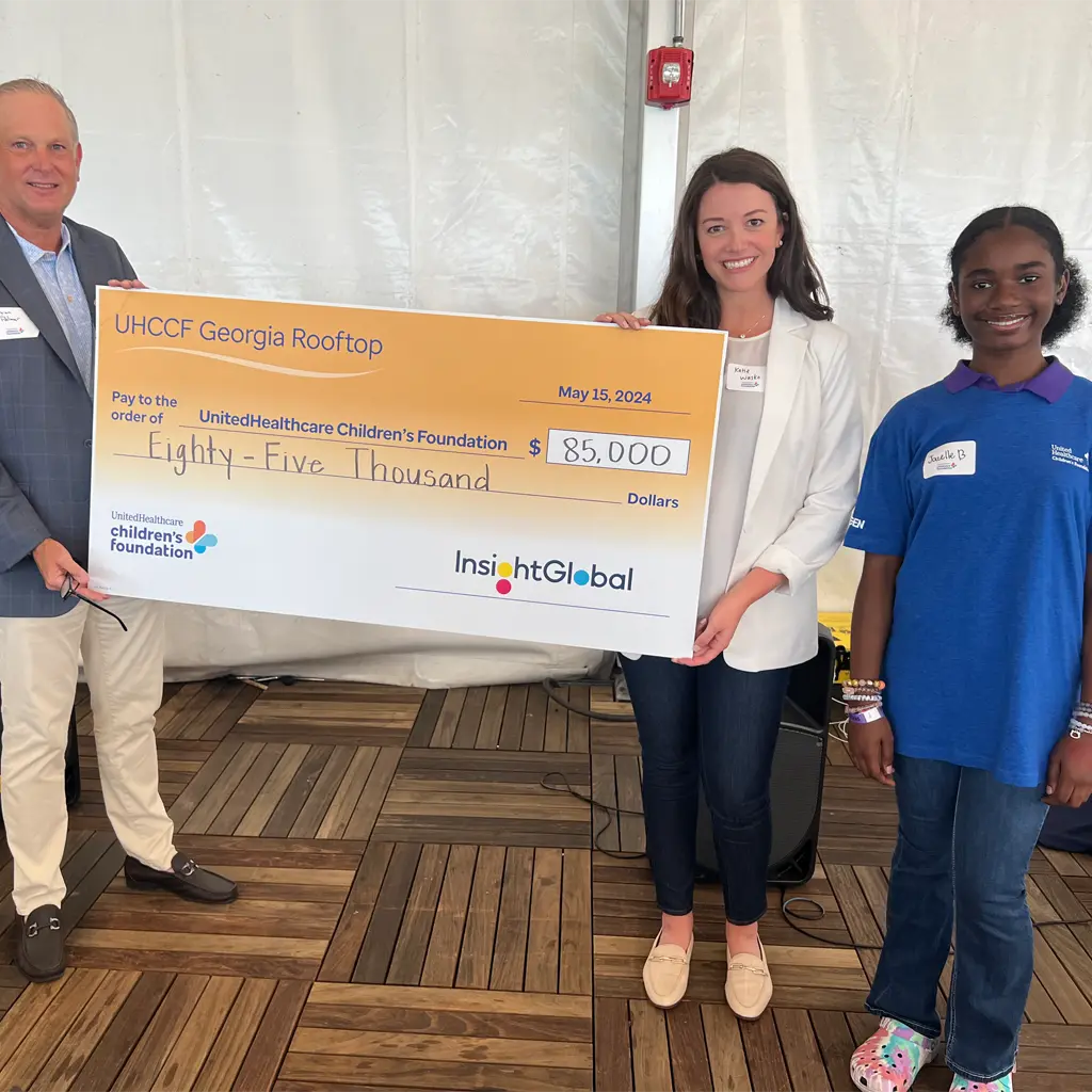 A man and woman hold a giant check for $85,000 between them, smiling at the camera. The check is from Georgia Rooftop and is made out to the UnitedHealthcare Children’s Foundation. A little girl in a blue UHCCF t-shirt stands next to the woman, smiling.