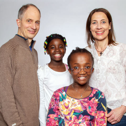 A family of four with a mom, dad, and two little girls stands together, smiling at the camera. The mom, Jill, has her hand on the little girl Maci’s arm. Maci is wearing glasses and standing closest to the camera.