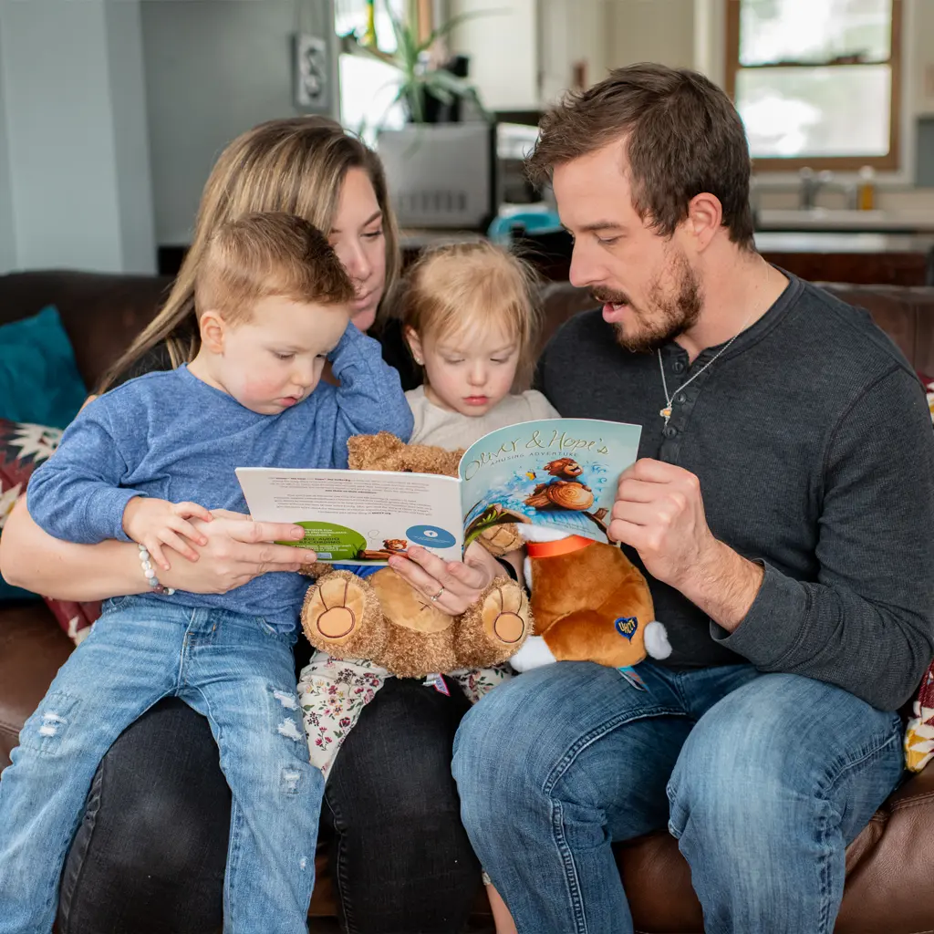 A family sits together on a couch, reading an Oliver and Hope book. The little boy sits on the mom’s lap, and she holds one side of the book open while the dad holds the other side and reads aloud. A little girl sits between them.