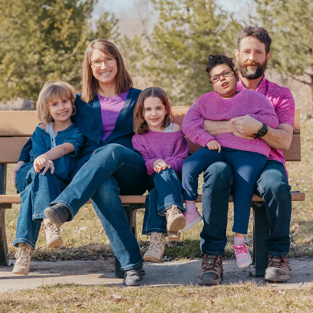 A family sits together on a bench. The dad is holding one of the little girls in his lap while the mom hugs the other two children on either side of her.