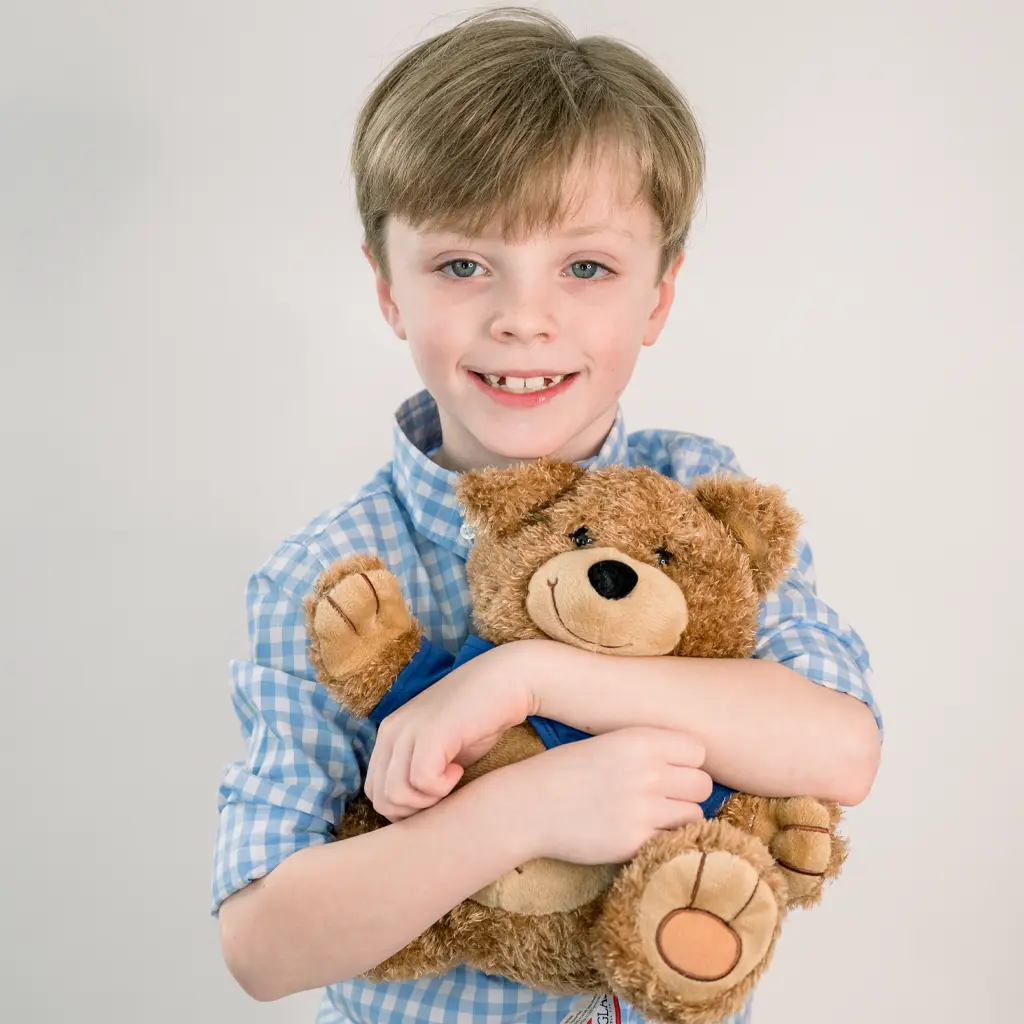 I little boy named Liam smiles at the camera, hugging an Oliver the Bear stuffed animal tightly to his chest.