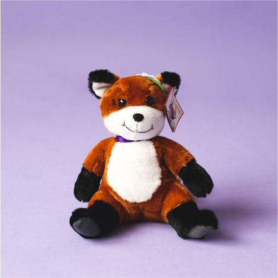 A Charlotte the fox stuffed animal wears a purple ribbon around her neck and sits on a light purple background.