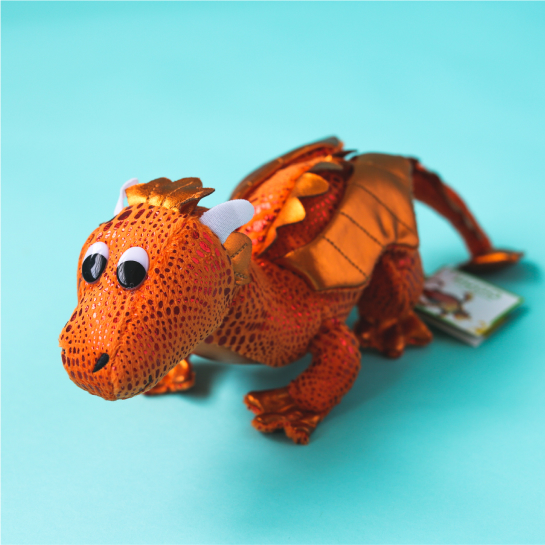 An orange Hugo the dragon stuffed animal with black spots and shiny metallic wings, tail, and fringe stands on a light turquoise background.