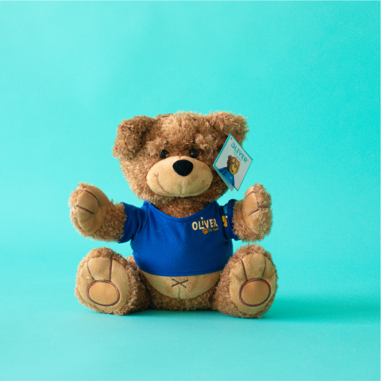 A stuffed Oliver the Bear wears a blue shirt with “Oliver” embroidered on the front and sits on a turquoise background.