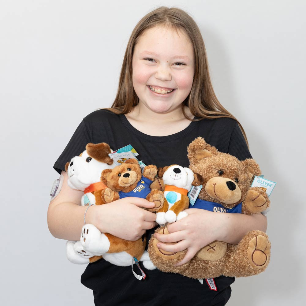 A little girl hugs an armful of stuffed animals, including Oliver the Bear, while smiling widely at the camera.